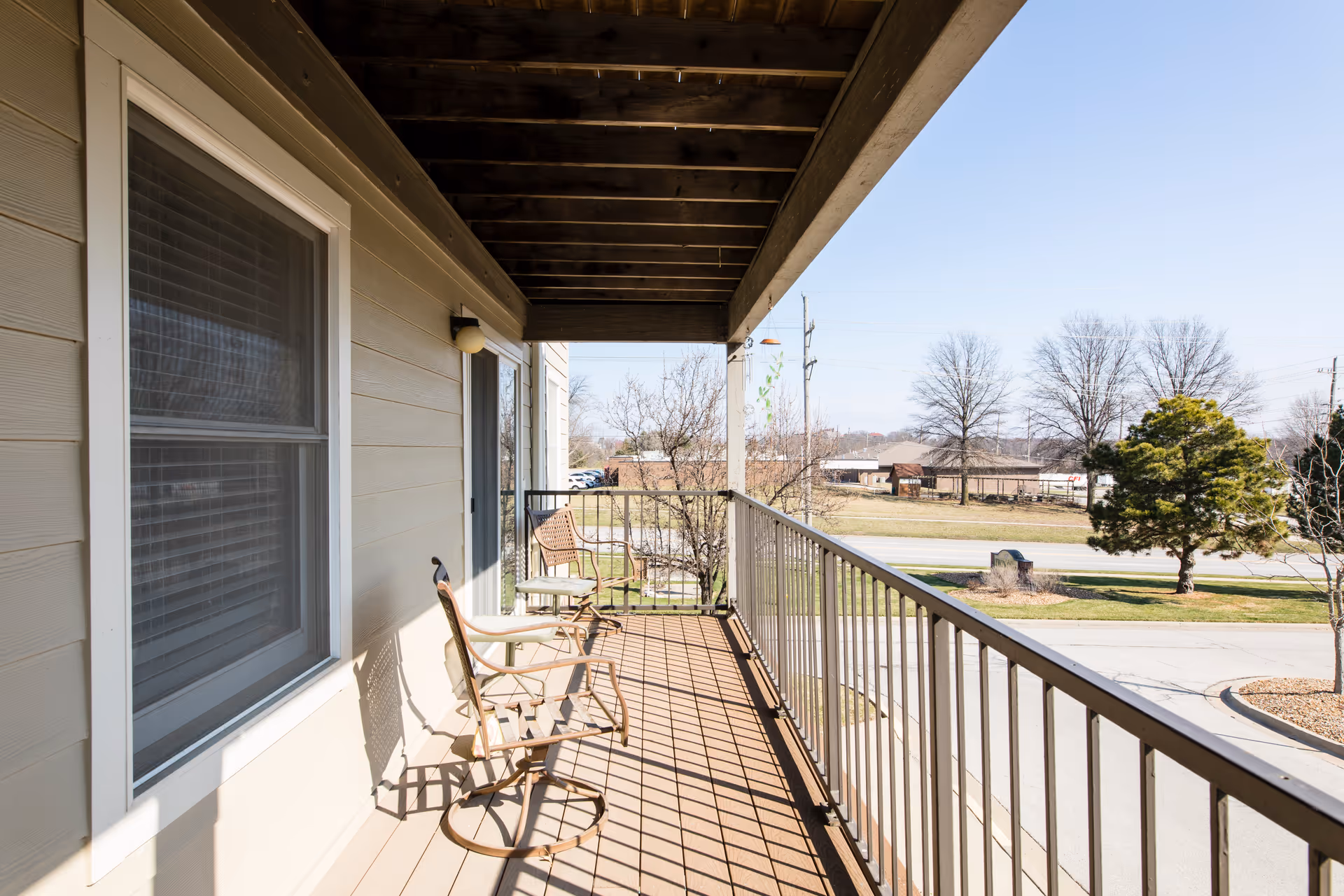 A balcony with two metal chairs and a small table, overlooking a street with trees and buildings in the distance under a clear blue sky.