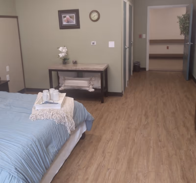 A bedroom with a bed covered in a light blue comforter and a beige throw blanket. On the bed is a tray with three white mugs and a book. Against the wall is a small table with a potted white orchid and a decorative pillow underneath. A framed photo and a clock hang on the light green wall. The room has wood flooring and an open doorway leading to another room with shelves and a plant on the door.
