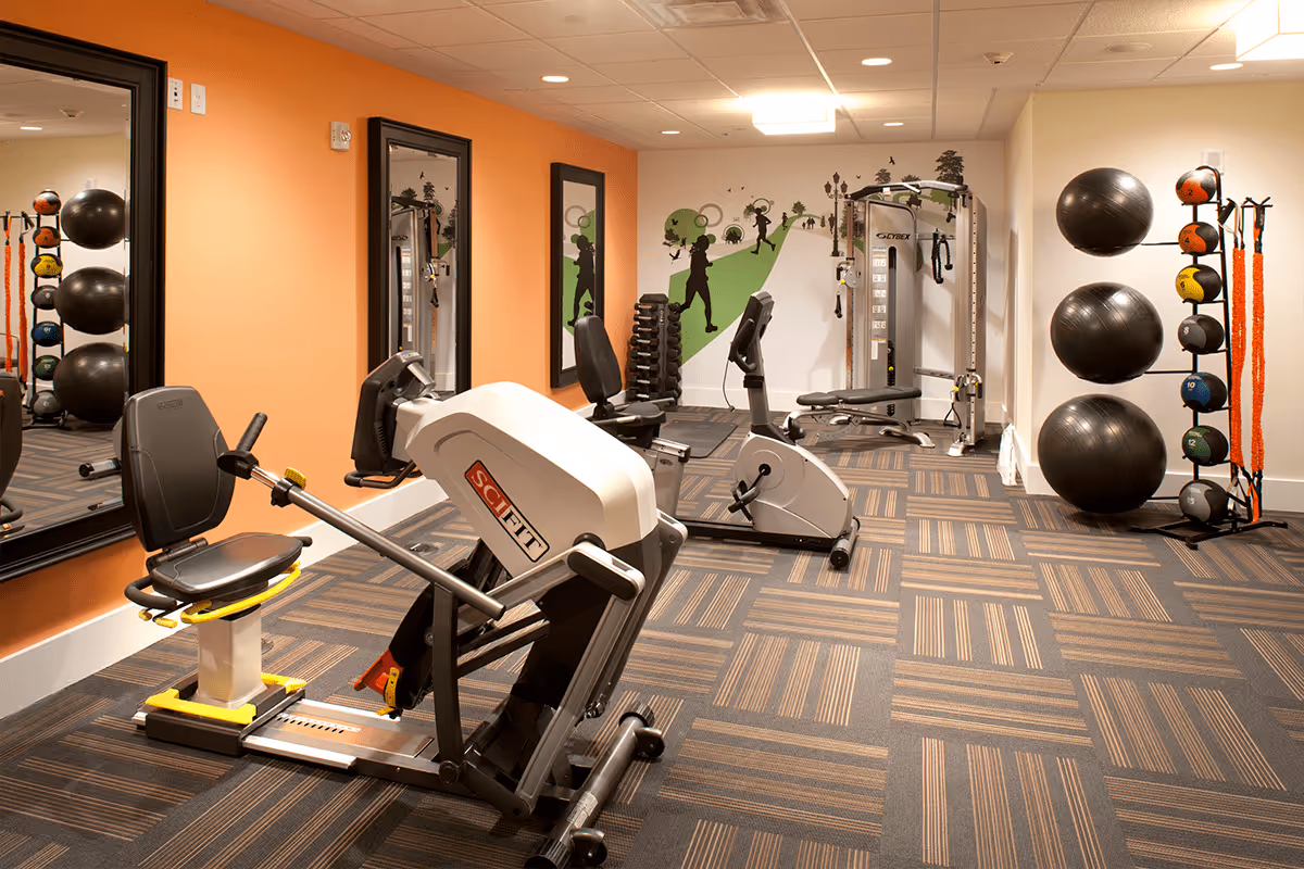 A well-lit fitness room with exercise equipment including a recumbent bike, a stationary bike, a multi-function weight machine, and racks holding medicine balls, stability balls, and resistance bands. The room has an orange accent wall with two large mirrors and a mural on the far wall depicting silhouettes of people walking and exercising in a park-like setting.