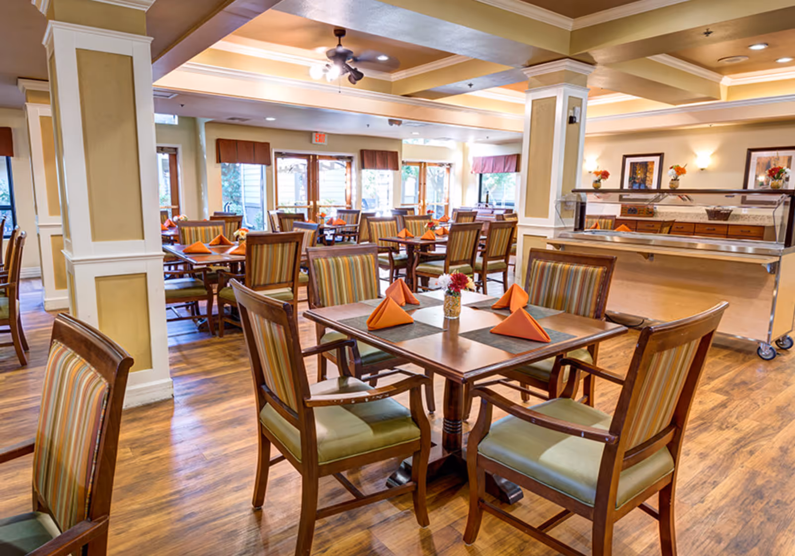 A bright and spacious dining room in a senior living facility with wooden tables and chairs. Each table is set with orange folded napkins and small flower arrangements. The room features large windows with natural light, wooden flooring, and a buffet serving area on the right side.