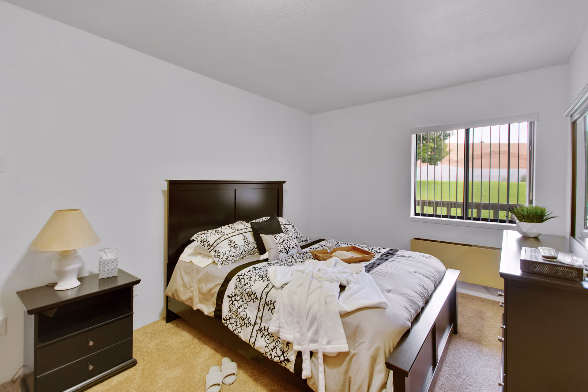A bedroom with a dark wooden bed frame and a neatly made bed with patterned bedding. A white robe and a tray with a glass and a book are placed on the bed. There is a nightstand with a lamp and a tissue box on the left side of the bed. A dresser with a plant and some items is on the right side near a window with vertical bars, showing a green lawn and a tree outside. The room has beige carpet and white walls.