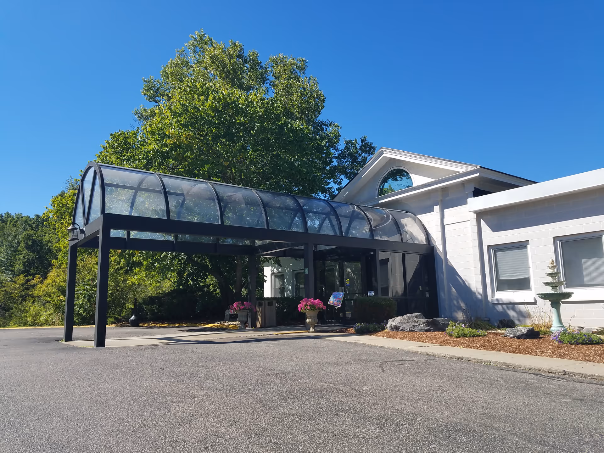 Front entrance of a white rehabilitation center with a covered glass canopy, potted flowers, a small fountain, and surrounding trees under a blue sky.