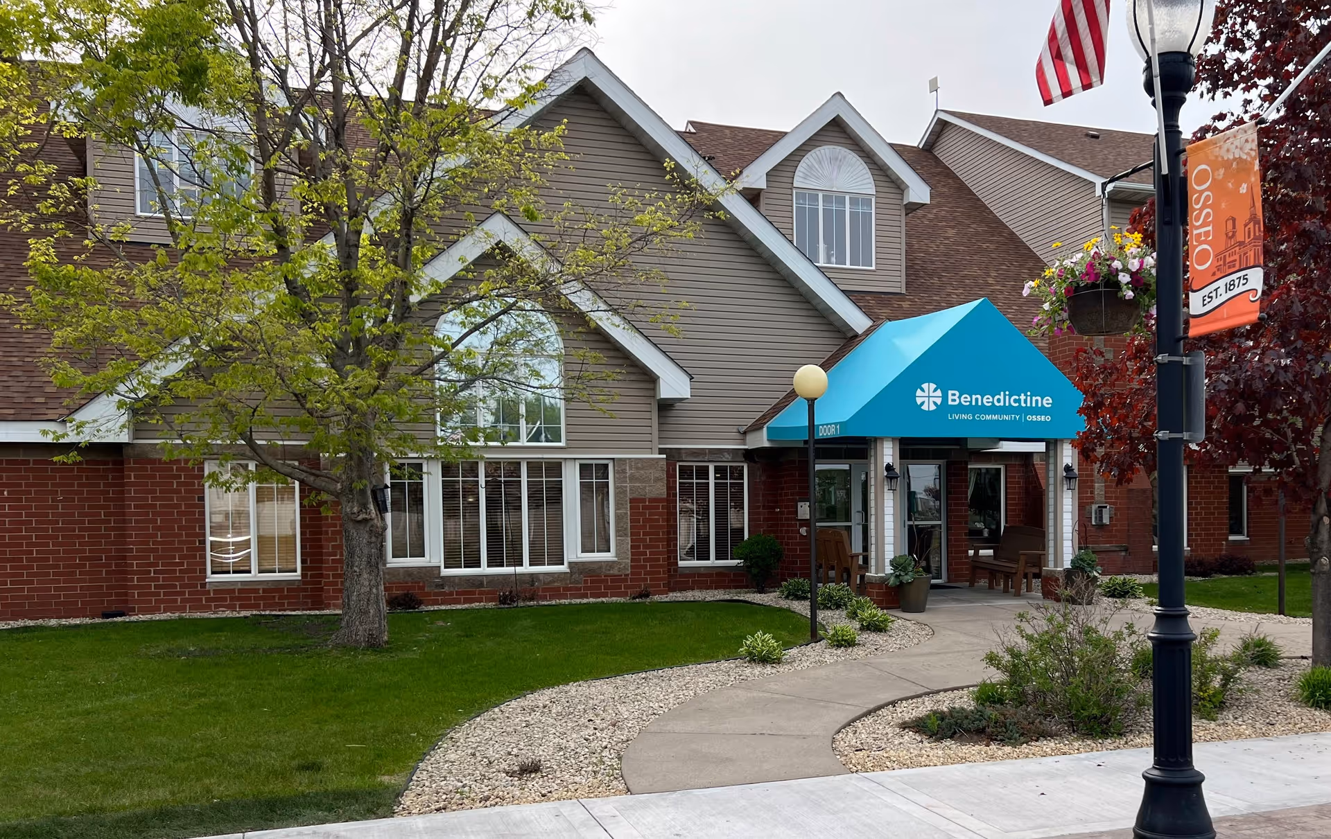 Front entrance of Benedictine Living Community-Osseo with a teal awning, walkway, landscaping and a street lamp with an Osseo banner.