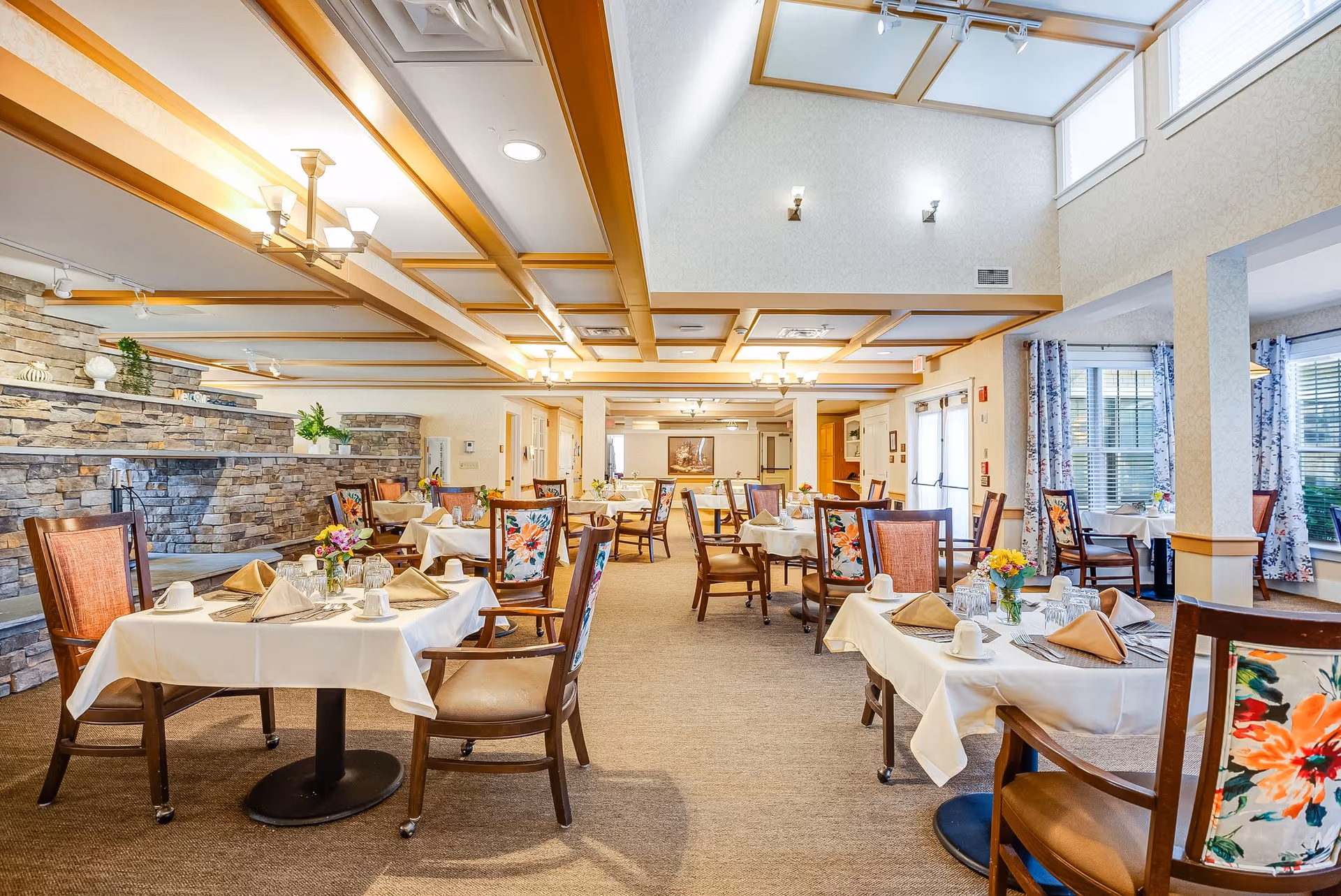 A bright and spacious dining room in a senior living facility with multiple tables covered in white tablecloths, each set with beige napkins, cups, and small flower arrangements. The room features wooden chairs with floral upholstery, a stone accent wall, large windows with floral curtains, and a ceiling with wooden beams and skylights allowing natural light to fill the space.
