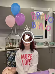 A woman with dark red hair wearing a white sweatshirt with the words 'MORE LOVE' in red letters sits in a room with gray cabinets and a window behind her. There are three balloons (blue, pink, and purple) attached to a decorative cage on the cabinet. Colorful text saying 'BRAVO BRAVO' is overlaid on the image.