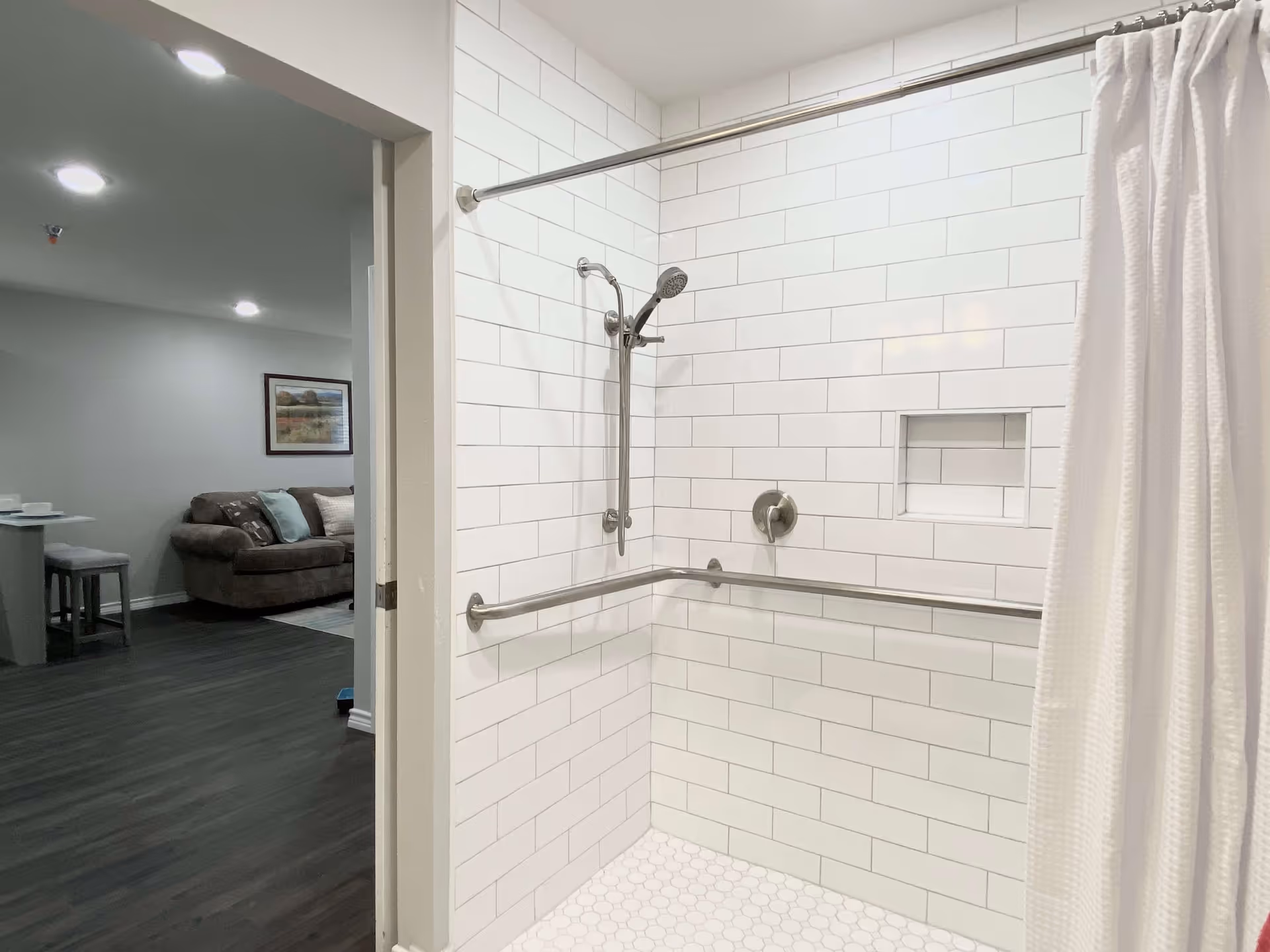 A white tiled shower area with a silver adjustable showerhead and a horizontal grab bar. The shower has a built-in niche for toiletries and a white shower curtain partially drawn to the side. Through the open doorway, a living room area with a brown couch, a framed picture on the wall, and a small dining area with stools is visible.