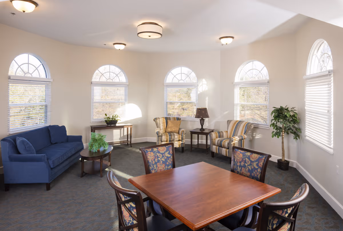 Bright communal living room with a blue sofa, striped armchairs, a wooden table with chairs, and arched windows letting in natural light.