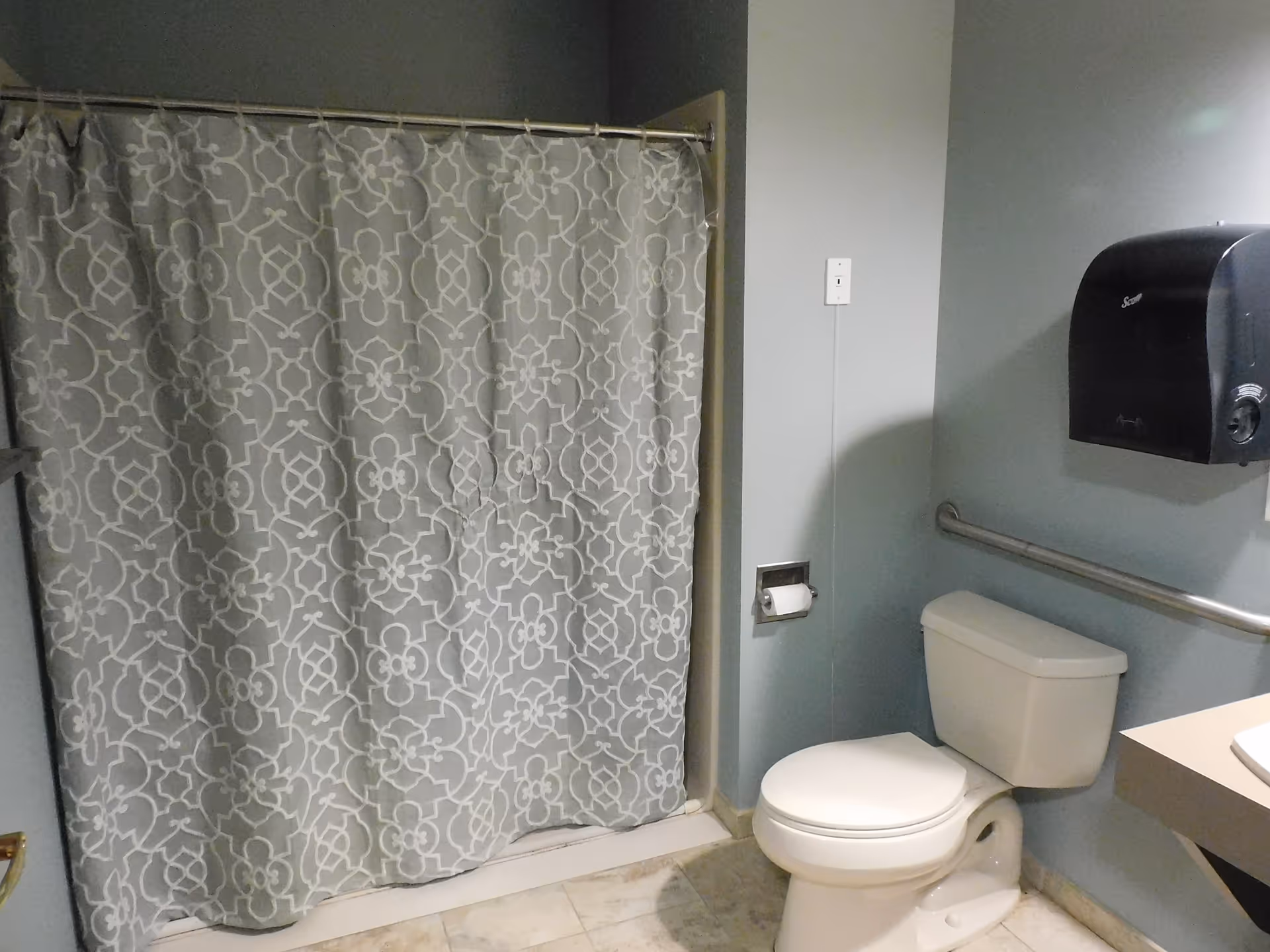 A bathroom with a white toilet, a gray patterned shower curtain, a wall-mounted paper towel dispenser, a grab bar next to the toilet, and a tiled floor.