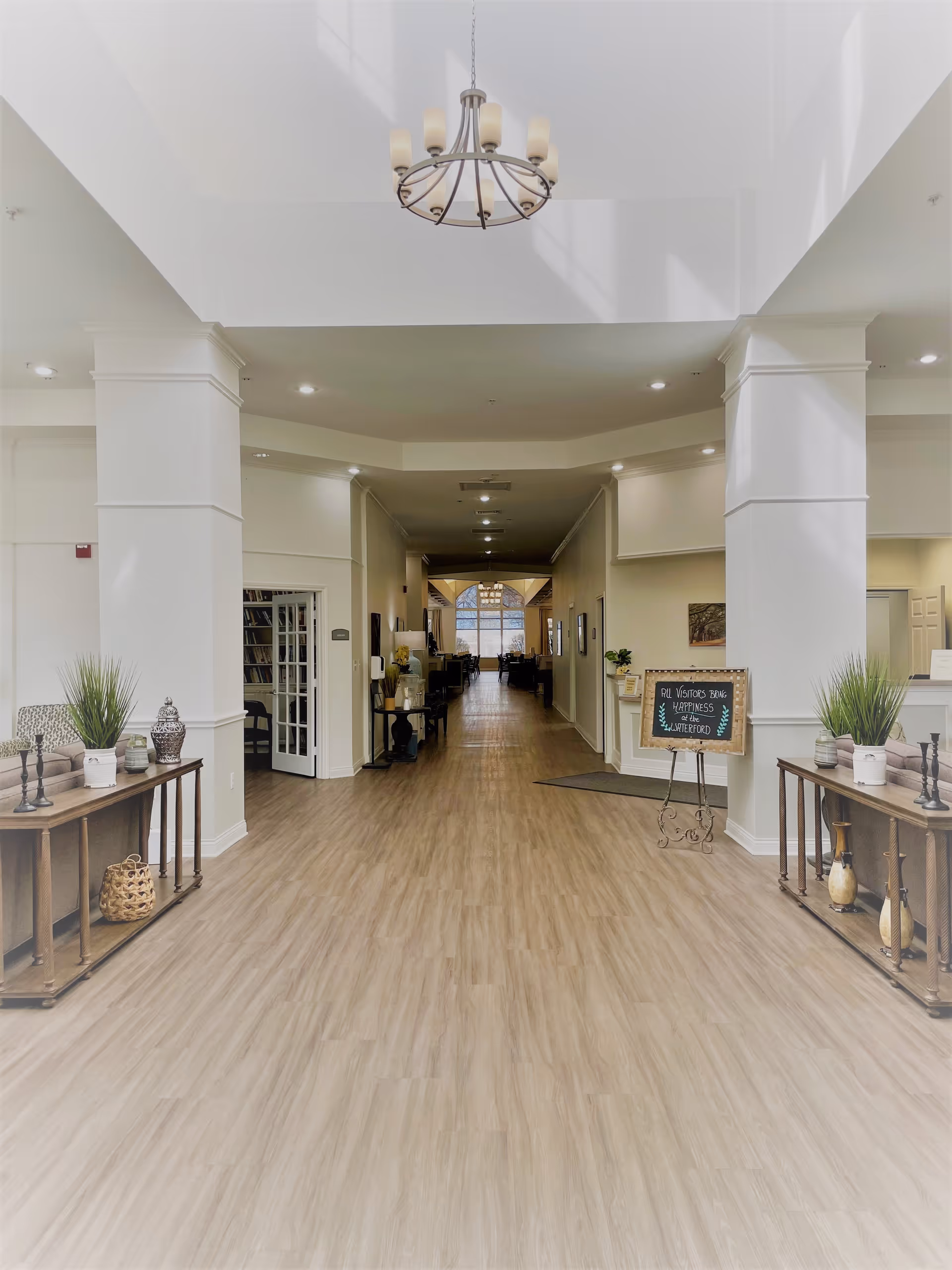 A spacious and well-lit hallway in a senior living facility with light wood flooring, white walls, and a chandelier hanging from the ceiling. There are decorative tables with plants and ornaments on either side of the hallway. A small chalkboard sign on an easel reads 'All visitors bring happiness to the Waterford.' The hallway extends into a seating area with large windows at the far end.