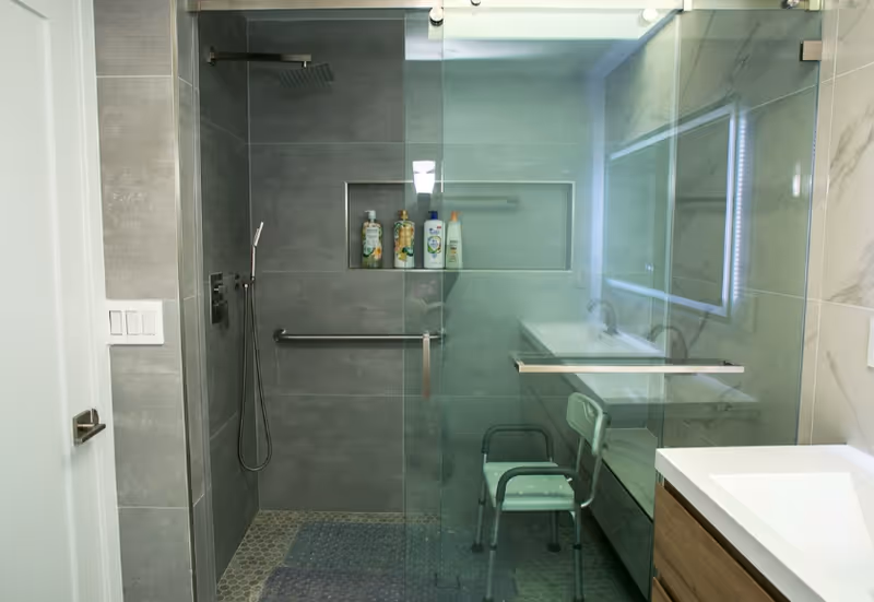 Modern glass-enclosed bathroom with shower, sink, built-in niche, and a small chair.