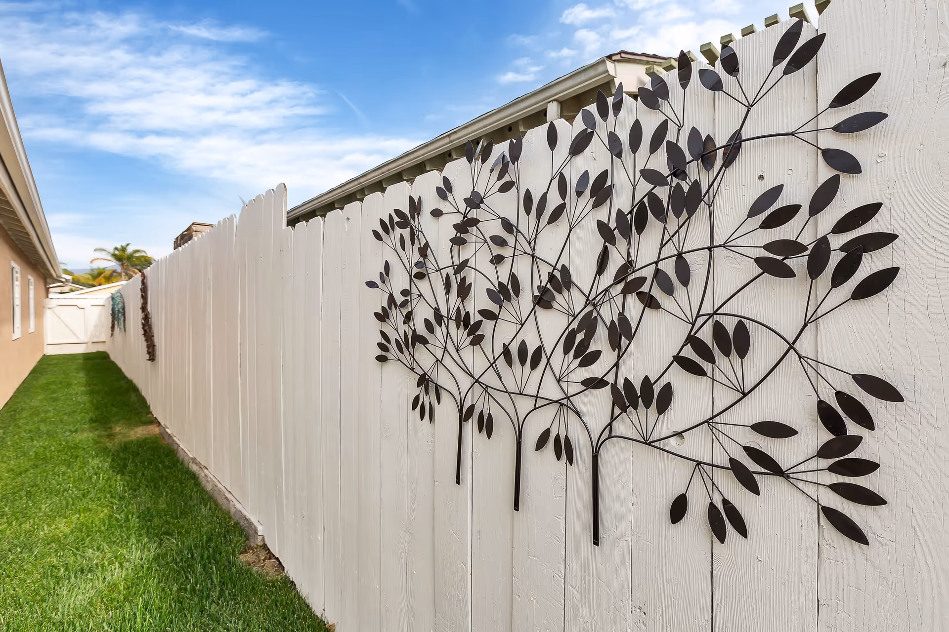Narrow grassy side yard with a white wooden fence featuring metal tree wall art beside a building under a blue sky.
