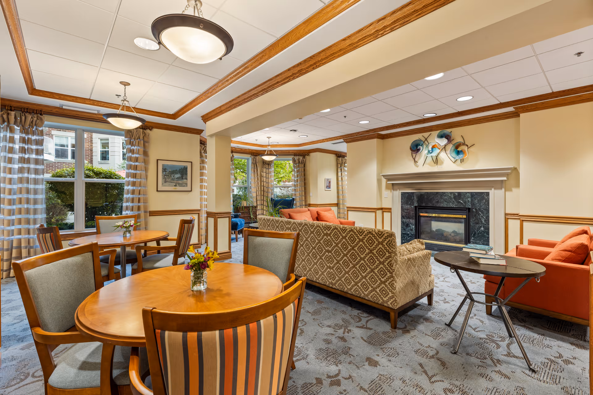 A cozy and well-lit common area in a senior living facility featuring round wooden tables with chairs, a seating area with patterned and orange sofas, a fireplace with decorative art above it, large windows with striped curtains, and ceiling lights.