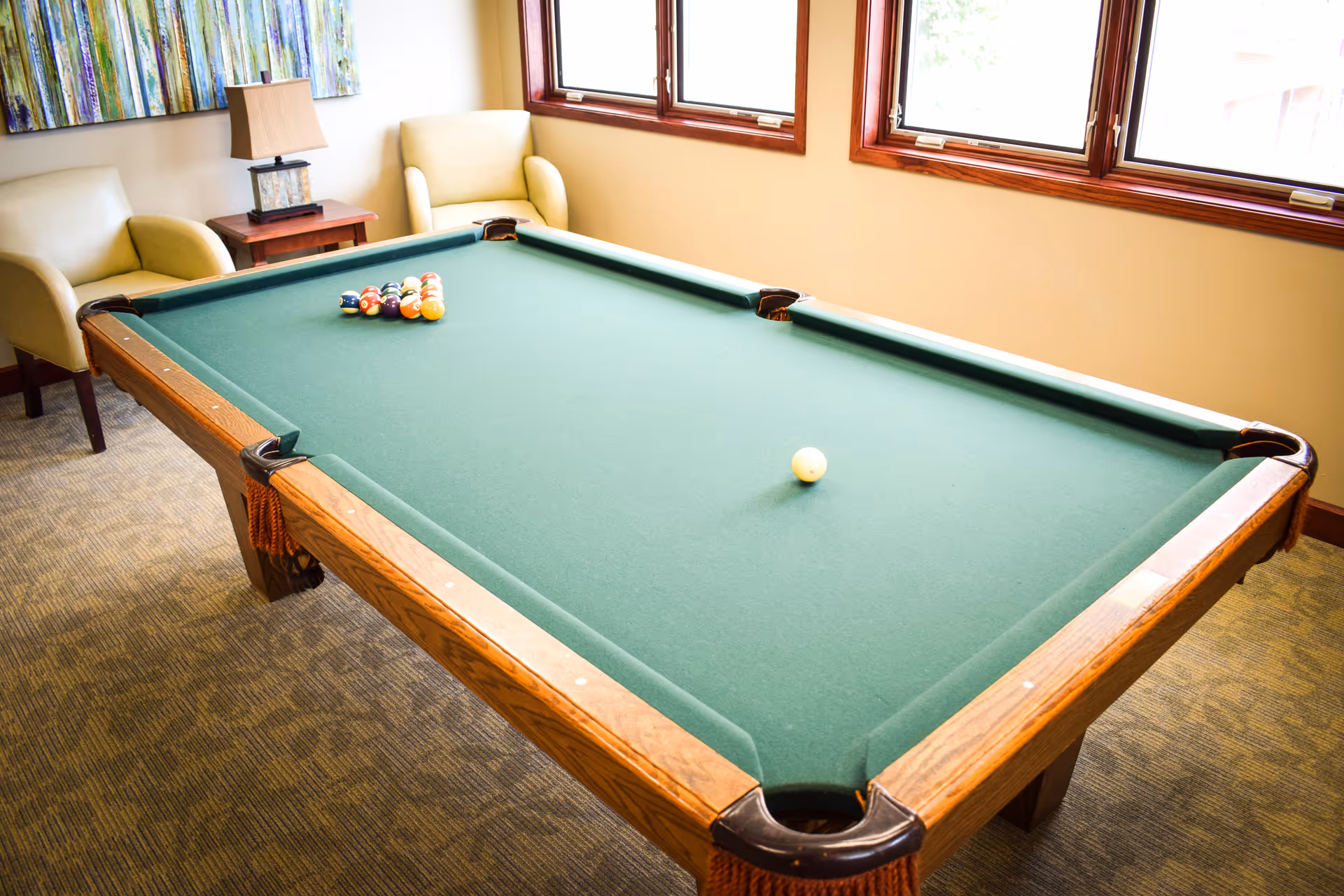 A billiards room with a green pool table set up for a game, two beige armchairs, a wooden side table with a lamp, and a colorful abstract painting on the wall. There are three windows letting in natural light.