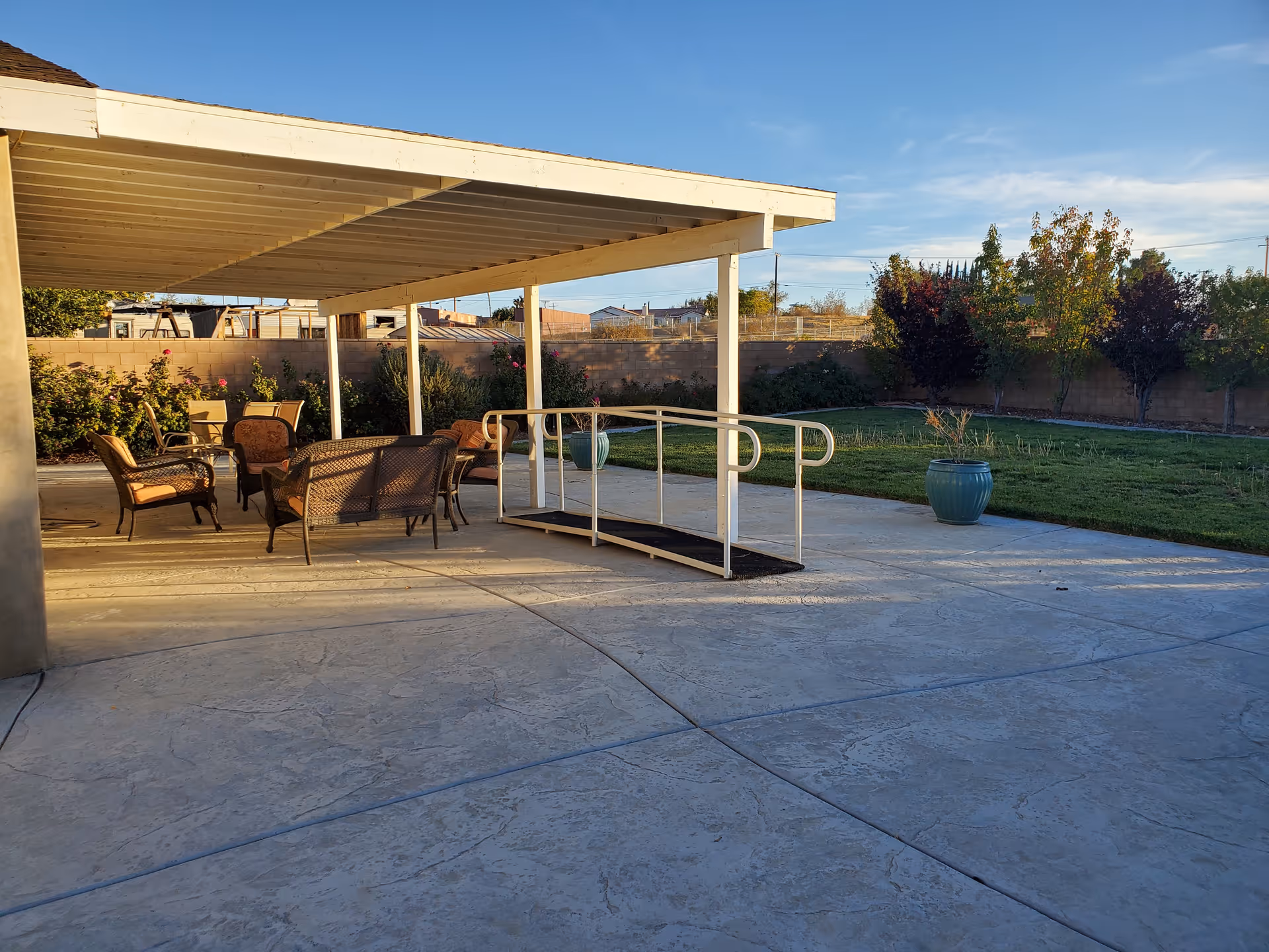 Outdoor patio area with a covered seating space featuring several cushioned chairs and a loveseat. There is a wheelchair-accessible ramp leading from the patio to a grassy yard with trees and plants along a brick wall fence under a clear blue sky.