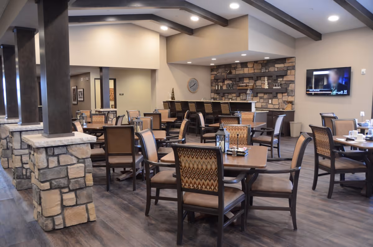 A spacious dining area with multiple wooden tables and chairs featuring patterned upholstery. The room has stone pillars, a stone accent wall with shelves, and a bar counter with stools. A wall-mounted TV is visible on the right side, and the ceiling has exposed dark wooden beams with recessed lighting.