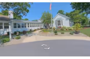 Exterior view of a single-story nursing center building with white siding and multiple windows. There is a paved driveway and landscaped garden beds with shrubs and flowers in front. An American flag is mounted on a pole near the entrance. The sky is clear and blue.