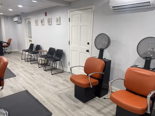 Interior view of a salon area with two orange salon chairs facing hair dryer stations, black chairs lined up against a white wall, a white door, and light wood flooring.