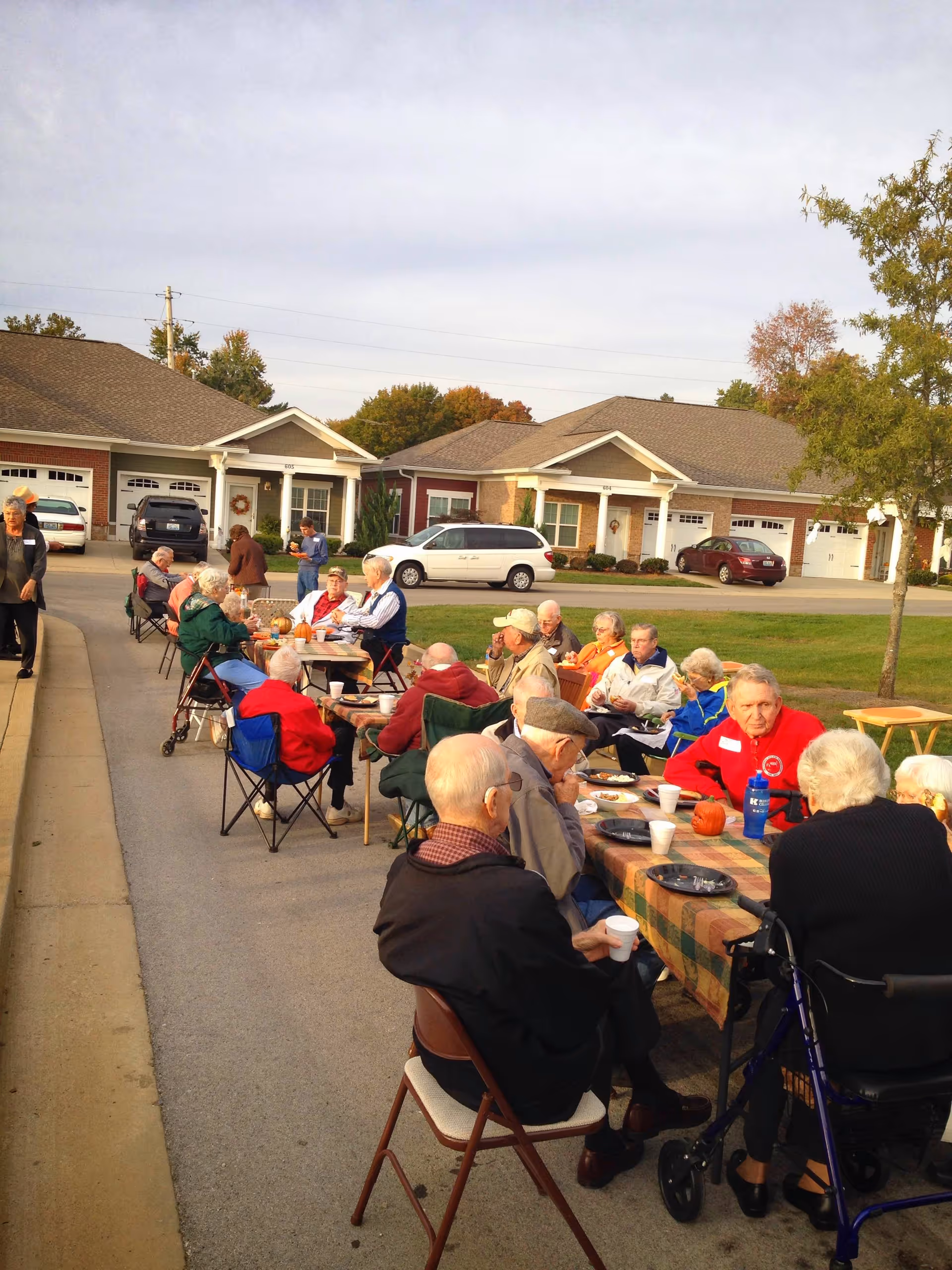 A group of elderly people sitting outdoors at tables covered with checkered tablecloths, enjoying a meal and socializing on a paved area in front of residential buildings with garages. Some people are seated in folding chairs, and there are small pumpkins on the tables, suggesting a fall or autumn gathering.