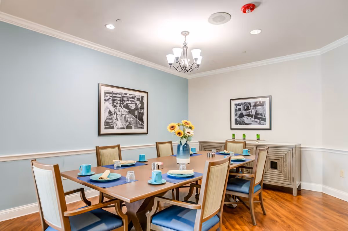 A dining room with a wooden table set for six people. The table has blue placemats, turquoise cups and plates, and beige napkins. A vase with sunflowers is placed in the center of the table. The room has light blue and white walls with two black and white framed photographs hanging. There is a wooden sideboard against the wall with three green glasses on top. The floor is wooden, and a chandelier with five lights hangs from the ceiling.