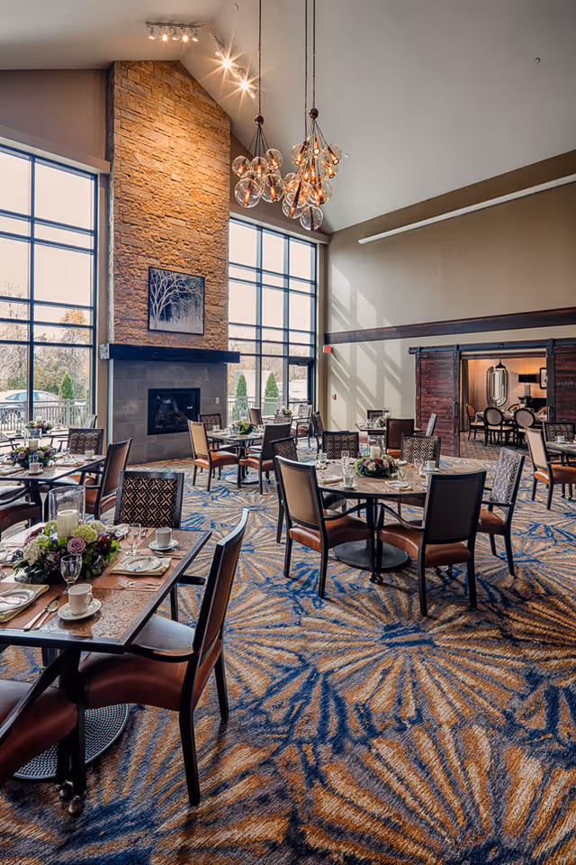 A spacious dining room with multiple tables set for meals, featuring chairs with brown cushions, floral centerpieces, and large windows letting in natural light. A tall stone fireplace with a framed artwork above it is visible, along with modern hanging light fixtures and a patterned carpet.