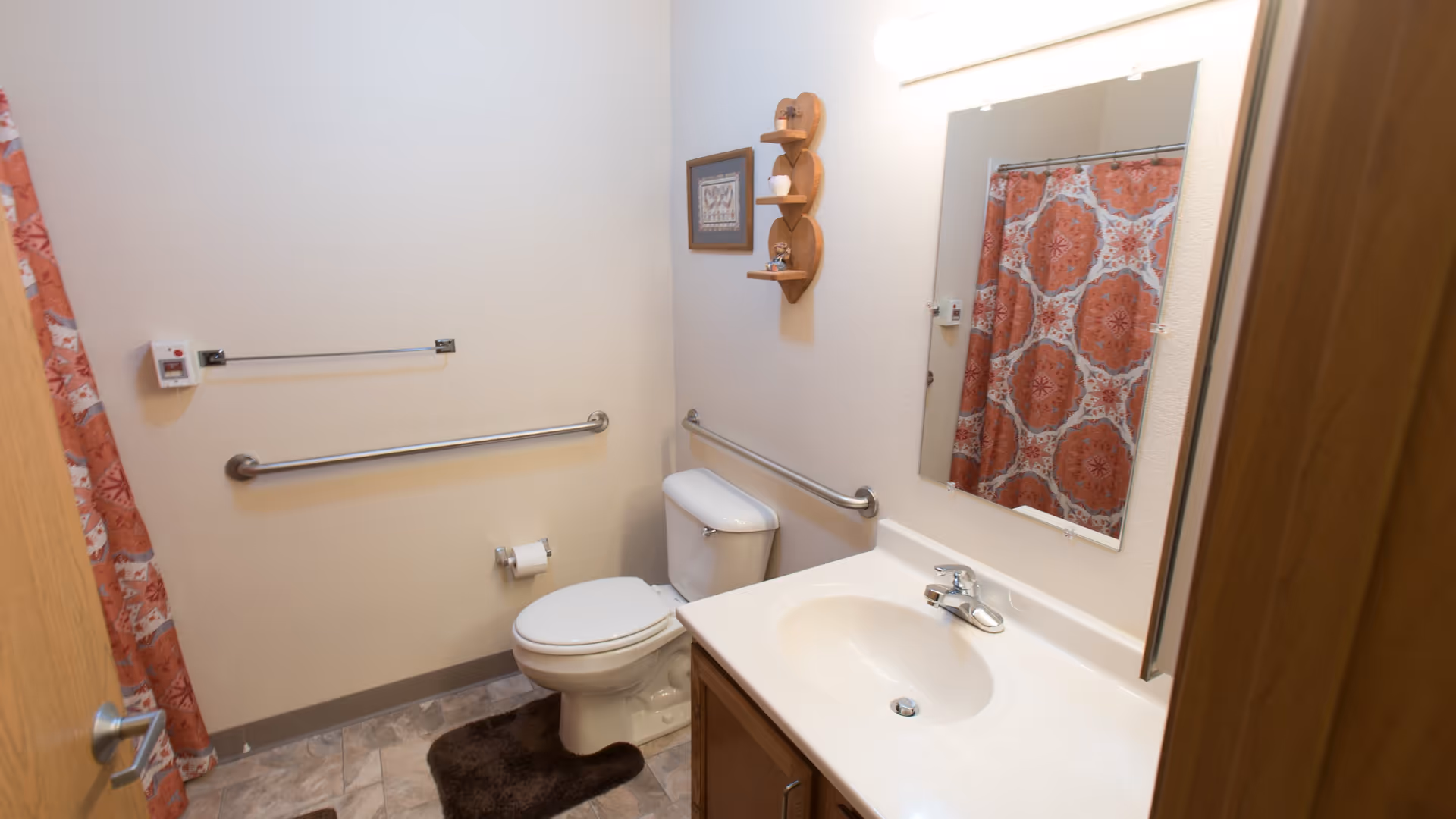 Small accessible bathroom with a toilet, sink vanity and mirror, grab bars, and a patterned shower curtain.