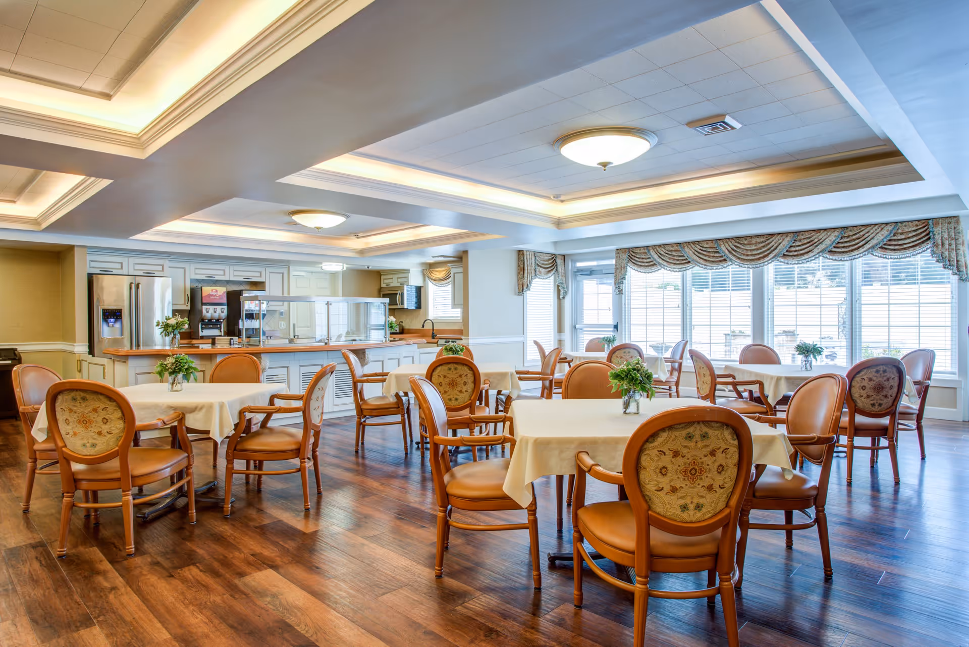 Bright dining room with round tables covered in white cloths, wooden chairs, a service counter, and large windows letting in daylight.