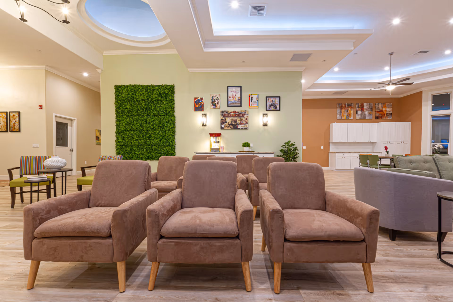 A spacious senior living common area with three brown armchairs in the foreground, a green wall decoration, framed artwork on a light green wall, and a popcorn machine. The room features a high ceiling with recessed lighting and a skylight. In the background, there are additional seating areas with chairs and sofas, a dining table, and white cabinetry along the far wall.
