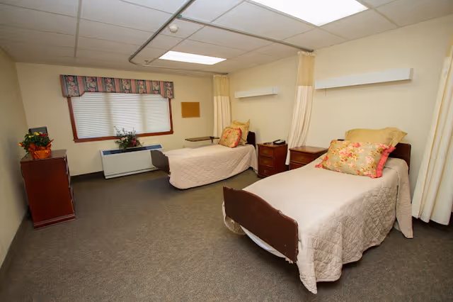 A simple, clean room with two single beds each covered with beige quilts and floral pillows. There are wooden nightstands between and beside the beds, a window with blinds and a floral valance, and a wooden dresser with a plant on top. The room has beige walls and carpeted floor, with ceiling lights and privacy curtains partially visible.