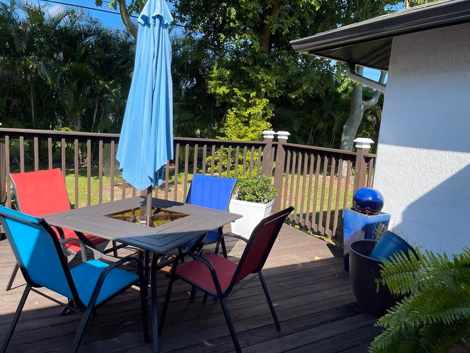 Outdoor wooden deck with a square table and four colorful chairs in red and blue. A closed blue umbrella is in the center of the table. The deck is surrounded by a wooden railing and lush green trees and plants are visible in the background. There are also some potted plants and a blue decorative sphere on a pedestal near the white exterior wall of a building.
