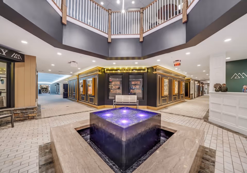 Interior view of a senior living facility lobby area with a modern square water fountain illuminated with purple lights in the center. Surrounding the fountain is a tiled floor and a two-story ceiling with a balcony railing above. In the background, there is a theater entrance with signs that say 'Movie Now Playing' and decorative gold trim. The area is well-lit with recessed lighting and has a clean, contemporary design.
