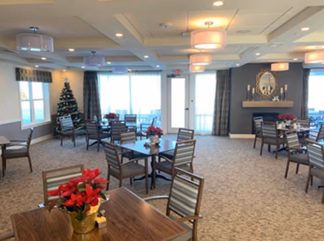 A spacious dining room with multiple wooden tables and chairs arranged neatly. Each table has a small red poinsettia plant as decoration. Large windows and glass doors allow natural light to fill the room. A decorated Christmas tree is visible in the corner near the windows. The room features modern ceiling lights, a fireplace with a round mirror above it, and curtains on the windows.