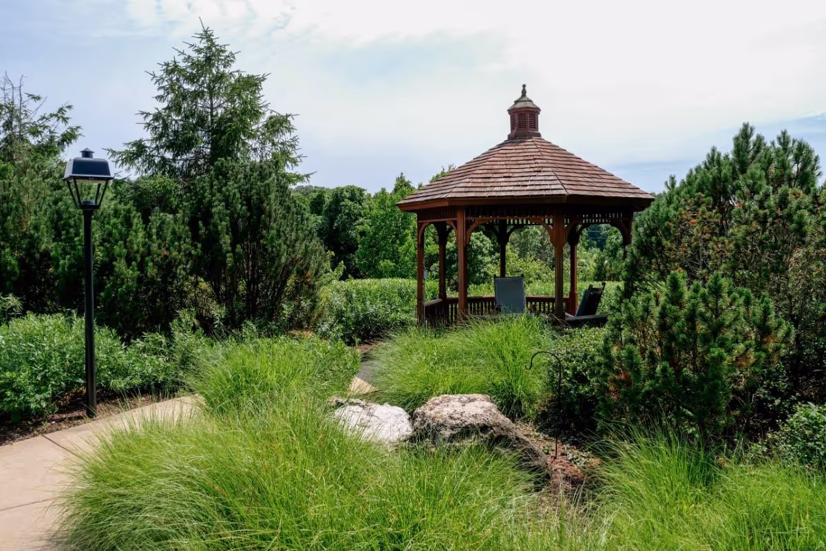 A wooden gazebo with chairs set among lush shrubs, tall grasses and a paved garden path with a lamppost.