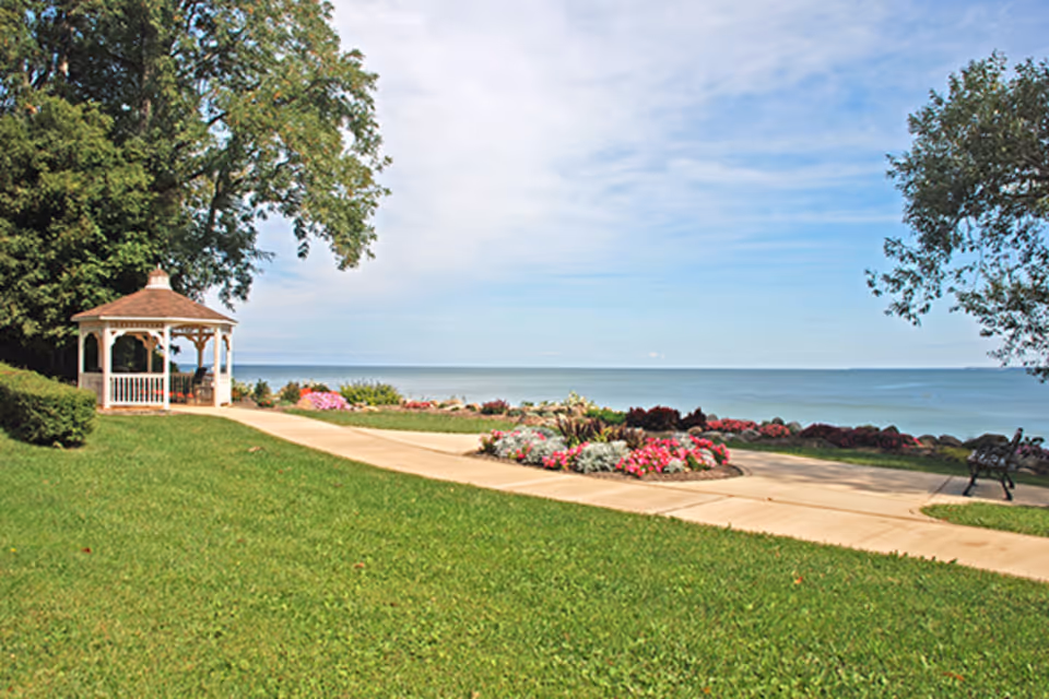 A lakeside garden with a white gazebo, manicured lawn, flowerbeds, a paved walkway, and a bench overlooking the water.