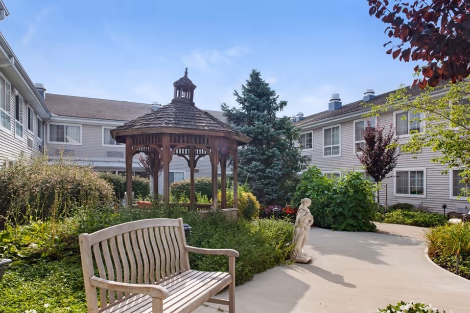 Outdoor garden area at Brighton Gardens of Edison featuring a wooden gazebo, a wooden bench, various shrubs and trees, a concrete pathway, and a small statue, with the facility building visible in the background under a clear blue sky.