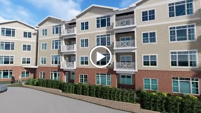 Exterior view of a multi-story senior living facility building with beige siding and red brick on the lower level, featuring multiple windows and balconies under a blue sky with some clouds.