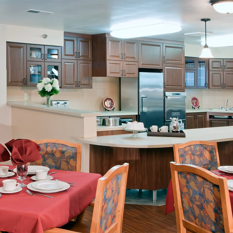 A dining area with tables covered in red tablecloths and set with white plates, cups, and silverware. Behind the dining area is a kitchen with wooden cabinets, a stainless steel refrigerator, oven, and a countertop with a cake on a stand, a coffee pot, and cups. A vase with white flowers is placed on the counter.
