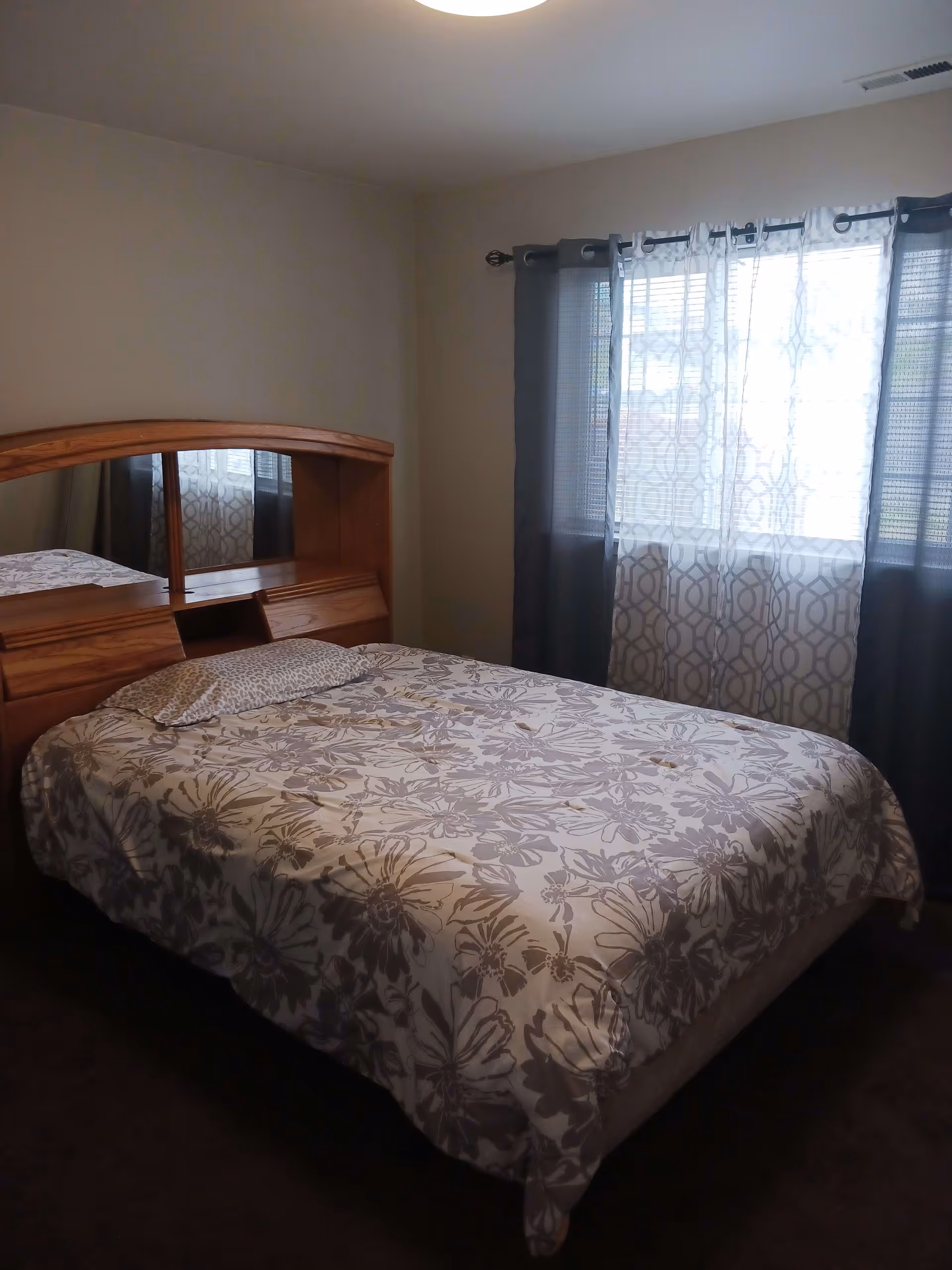 A bedroom with a bed covered in a floral patterned bedspread and a matching pillow. The bed has a wooden headboard with built-in shelves and mirrors. There is a window with sheer and dark curtains allowing natural light into the room.