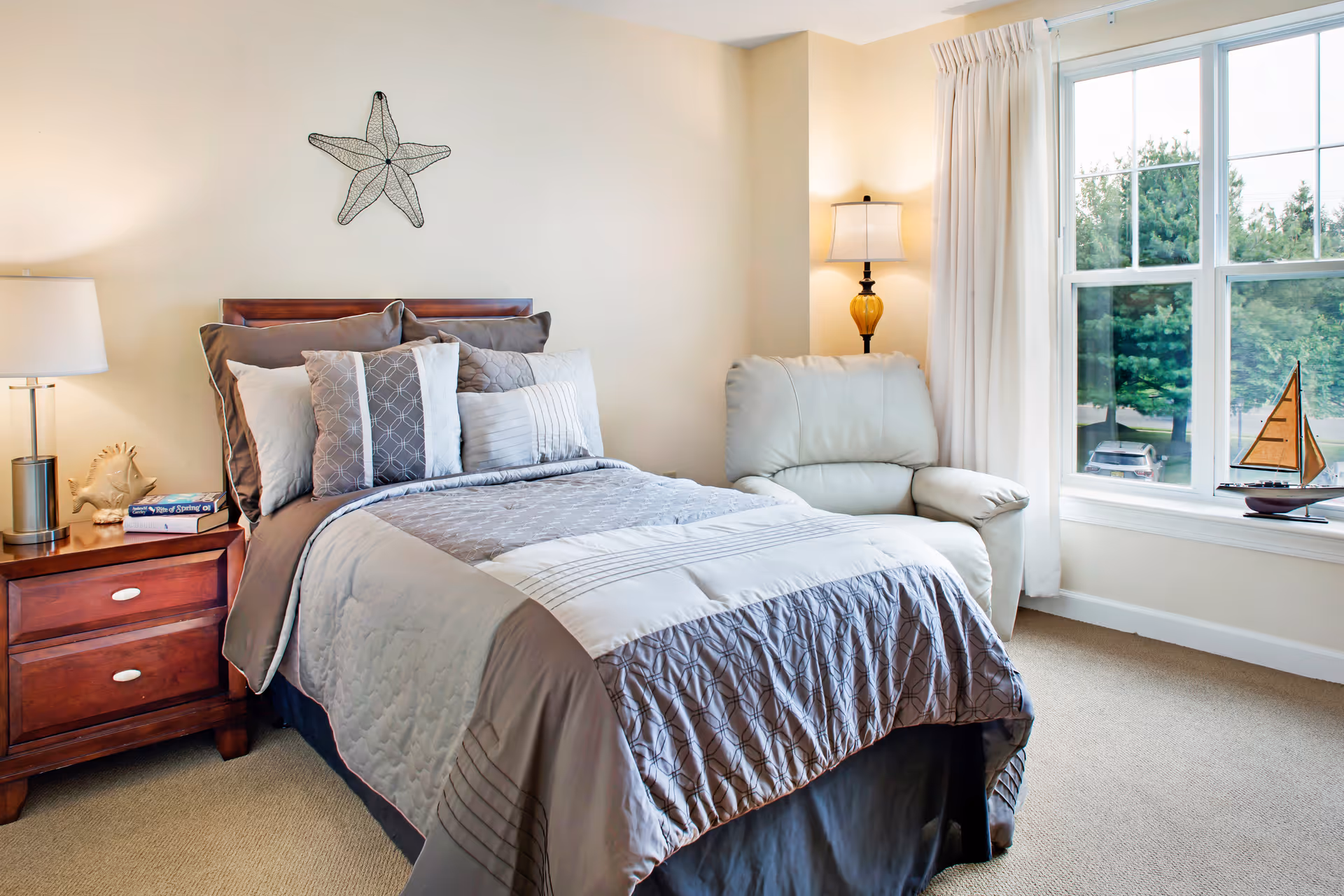A neatly made bed with multiple pillows and a gray patterned comforter in a bright bedroom. Next to the bed is a wooden nightstand with a lamp, a decorative seashell, and a book. A comfortable armchair is placed near a large window with white curtains, and a small decorative sailboat is on the windowsill. The walls are light-colored with a starfish wall decoration above the bed.