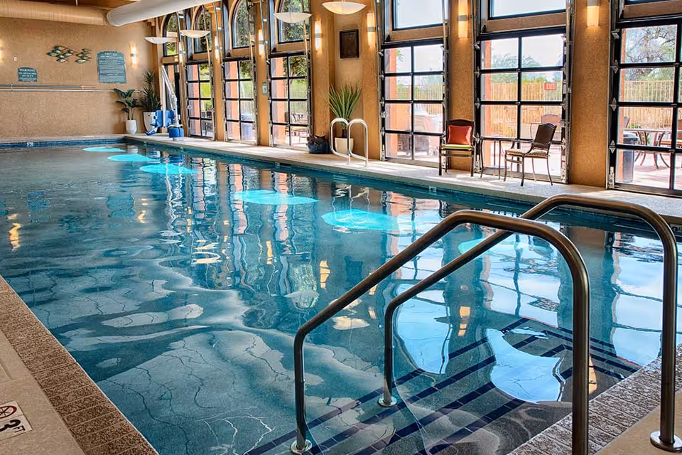 Indoor swimming pool with metal handrails, seating, and large glass doors along one side.