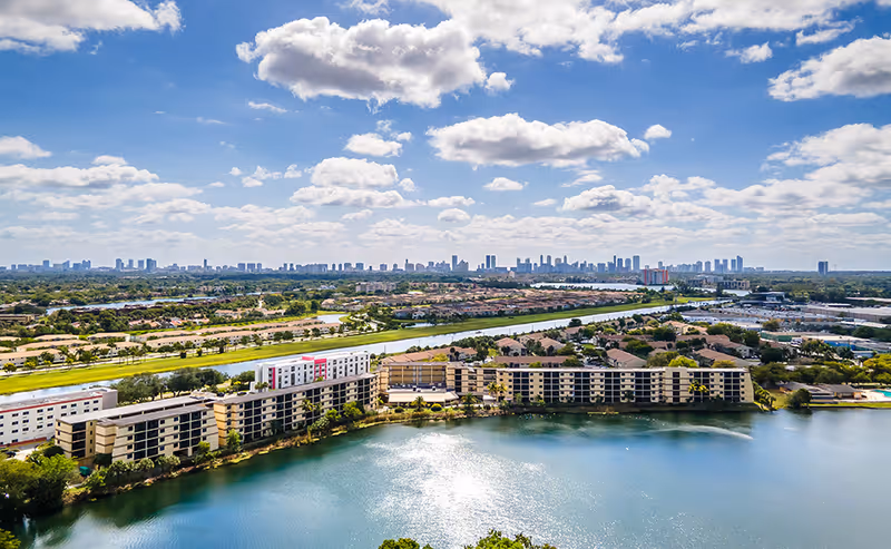 Aerial view of a lakeside apartment complex with several mid-rise buildings along a lake and a distant city skyline under a partly cloudy sky.