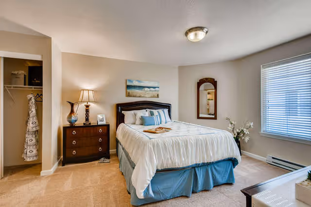 A bright and cozy bedroom with a neatly made bed featuring white and blue bedding. There is a wooden nightstand with a lamp and decorative items next to the bed. A mirror hangs on the wall beside a window with blinds, and an open closet with hanging clothes is visible on the left side. The room has beige walls and carpeted flooring.