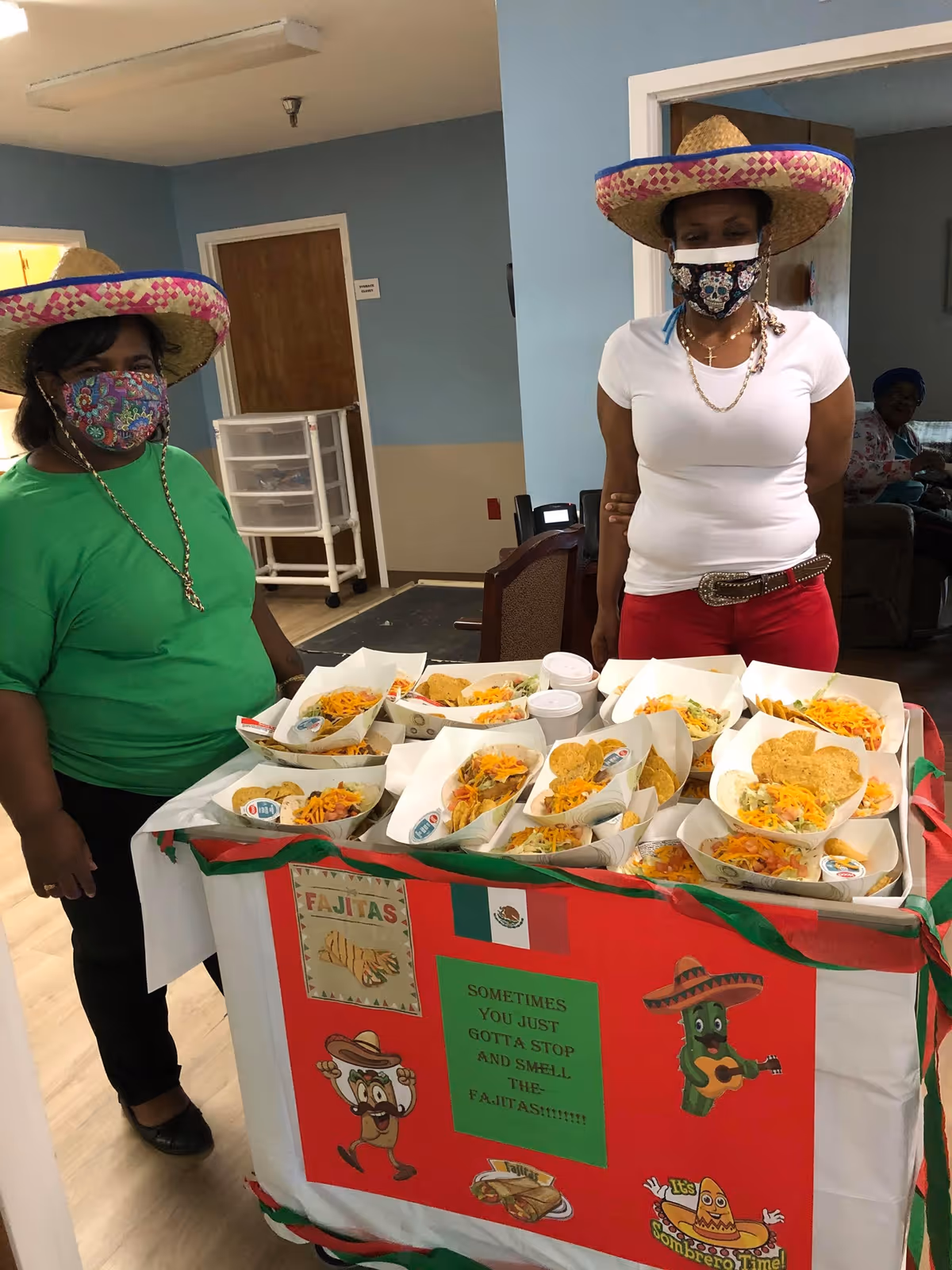 Two women wearing sombreros and face masks stand behind a table filled with trays of food, likely tacos with chips and cheese. The table is decorated with a red cloth featuring Mexican-themed images and a sign that reads, 'Sometimes you just gotta stop and smell the fajitas.' The setting appears to be an indoor common area with blue and beige walls.