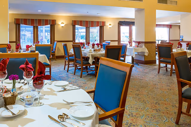 A dining room in a senior living facility with round tables set with white tablecloths, plates, silverware, wine glasses, and red folded napkins. The room has blue cushioned wooden chairs, patterned carpet, and windows with striped valances allowing natural light to enter.