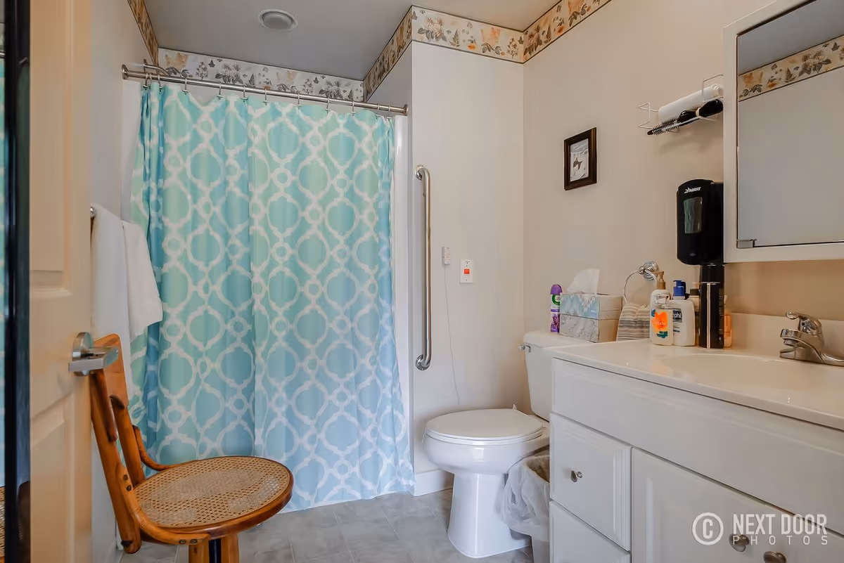 A bathroom with a white toilet, a white vanity with a sink, and a large mirror above it. There is a blue and white patterned shower curtain on a rod, a wooden chair with a woven seat, and a towel hanging on the wall. Various toiletries and a tissue box are placed on the vanity. The walls are light-colored with a floral border near the ceiling.