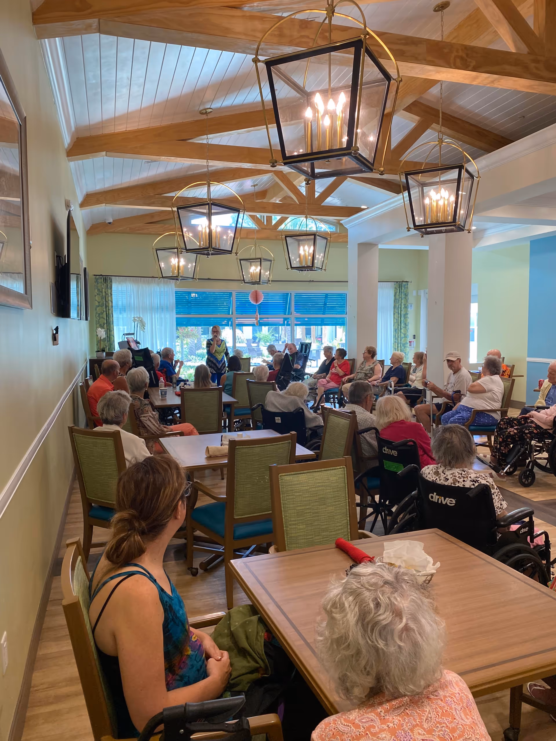 A group of elderly people seated in a large, well-lit room with wooden beams on the ceiling and hanging lantern-style lights. Some individuals are in wheelchairs while others sit on chairs around tables. A woman stands at the front of the room speaking to the group. Large windows with blue blinds and sheer curtains allow natural light to enter the space.