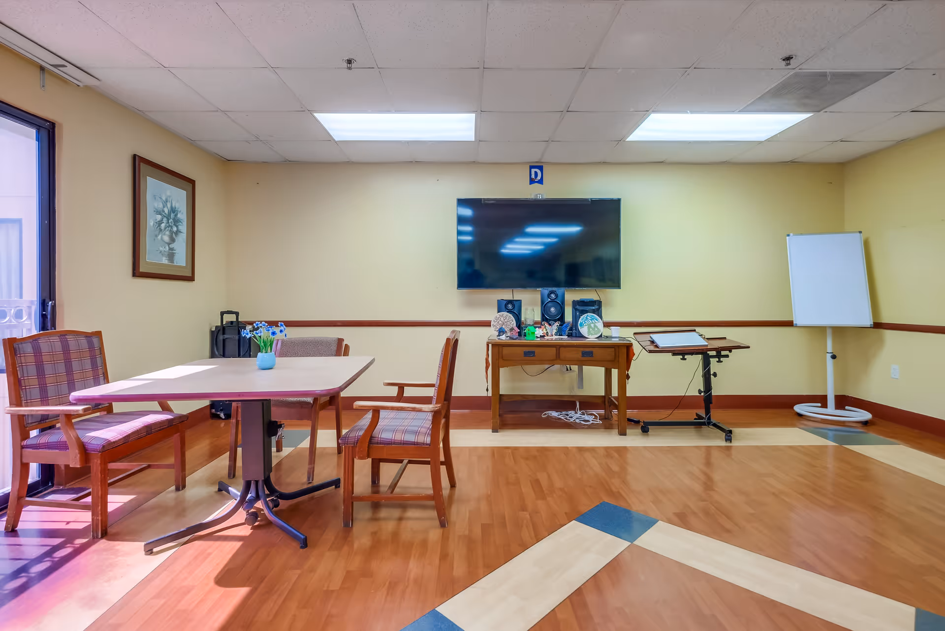 A small common area with a table and four plaid cushioned chairs, a flat-screen TV mounted on a beige wall, a wooden table with speakers and decorative items, a whiteboard on a stand, and a framed floral picture on the wall. The floor has a wood pattern with some colored tiles.