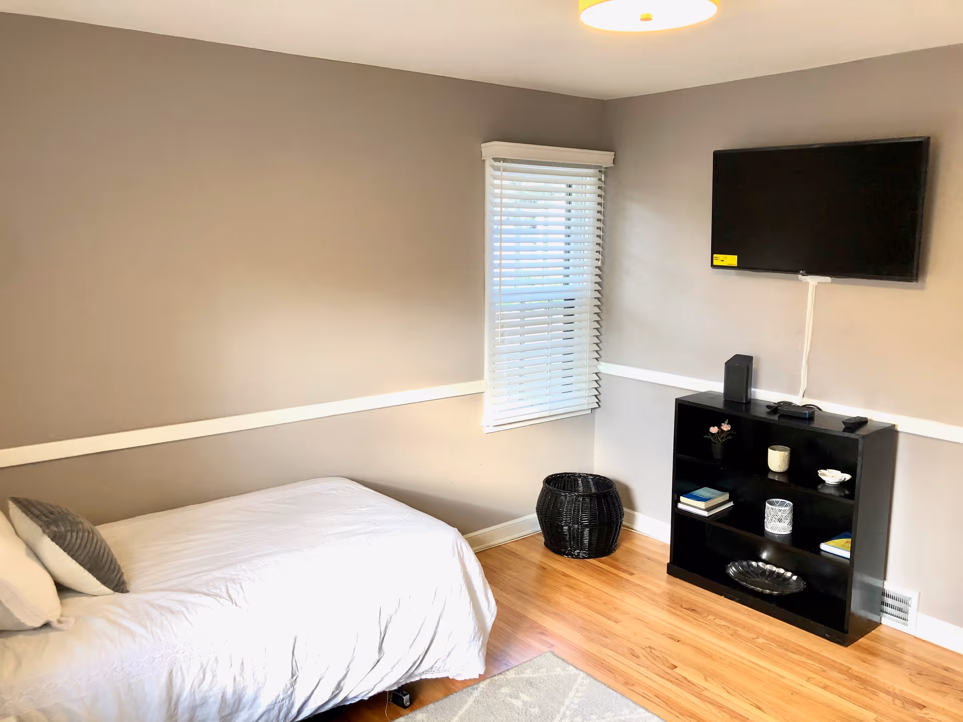 A simple bedroom with a single bed covered in white bedding and a gray pillow. There is a window with white blinds on the wall, a black woven basket on the floor, and a black shelving unit with books, decorative items, and a mounted flat-screen TV above it. The room has light brown walls and wooden flooring.