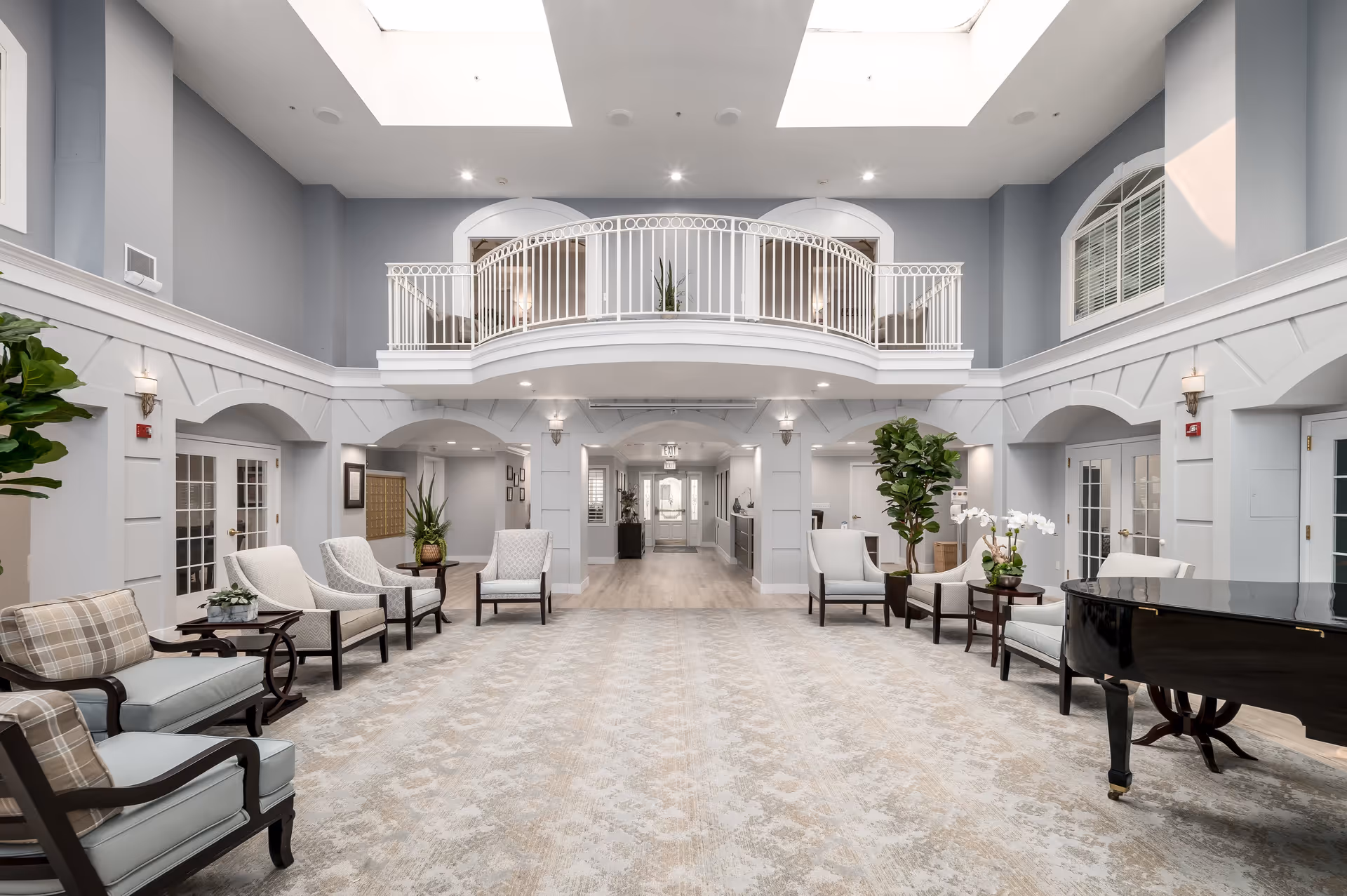 A spacious and elegant senior living facility common area with high ceilings and skylights. The room features a light gray and white color scheme, comfortable armchairs arranged around small tables, potted plants, and a black grand piano on the right side. There is a balcony with white railing overlooking the area and multiple doorways leading to other rooms.