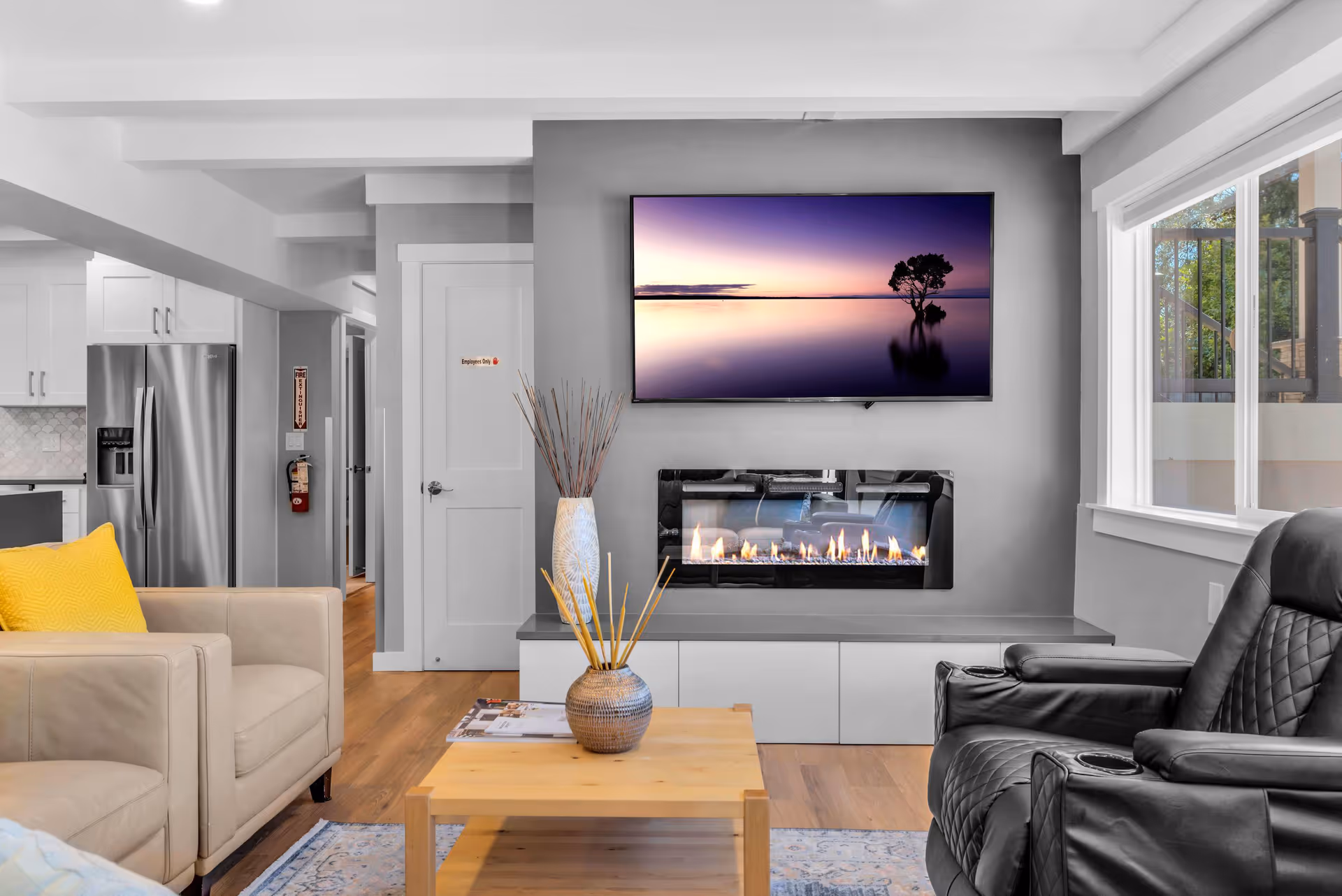 A modern living room featuring a wall-mounted TV above a linear fireplace, seating, and a coffee table with a view into the kitchen.