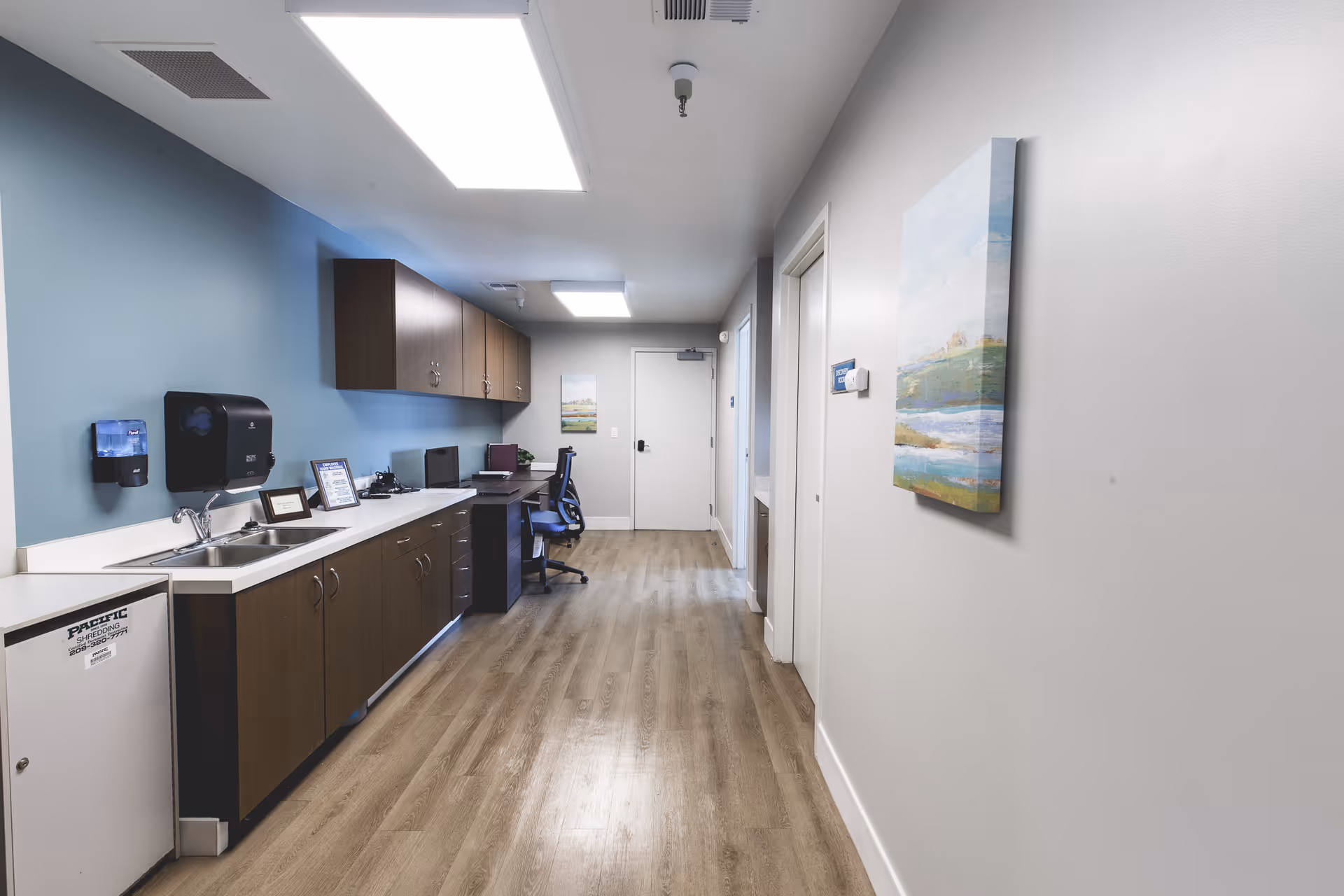 A clean, well-lit hallway in a senior living facility with wood flooring and light blue and white walls. On the left side, there is a countertop with a sink, cabinets, a small fridge, and office equipment including a computer and chair. On the right wall, there is a colorful landscape painting. The hallway leads to a closed white door at the end.